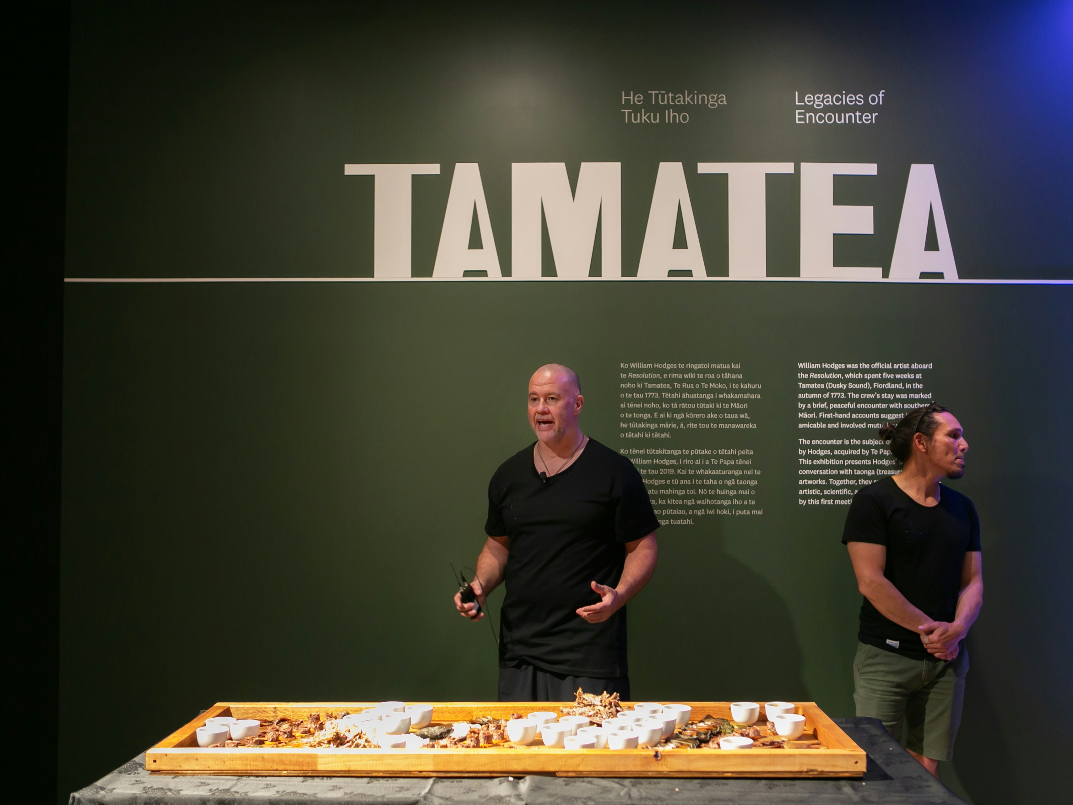 Simon Kaan and Ron Bull of Kaihaukai at Te Papa, 3 March 2020. Te Papa (150720) Two me stand against a green wall with the word “Tamatea” written on it, talking to a crowd behind the camera. In front of them is a table with an array of foods on it