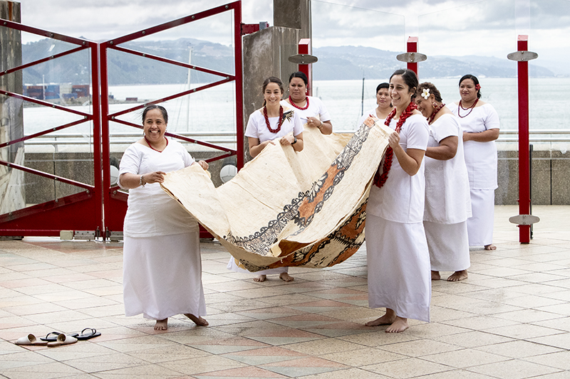 Seven women wearing white are carrying a large cloth made of bark cloth across a concourse