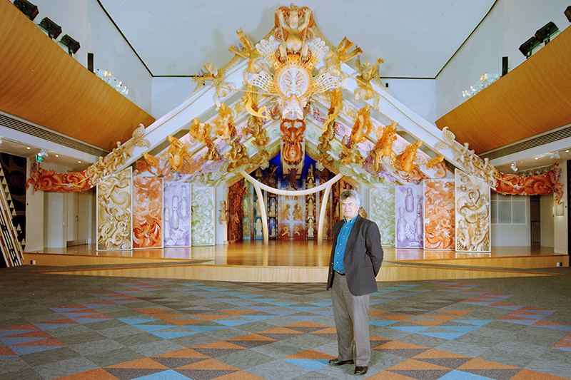 A man stands in a large room on patterned carpet. There's a stage behind him with a lot of colourful shapes made out of MDF.