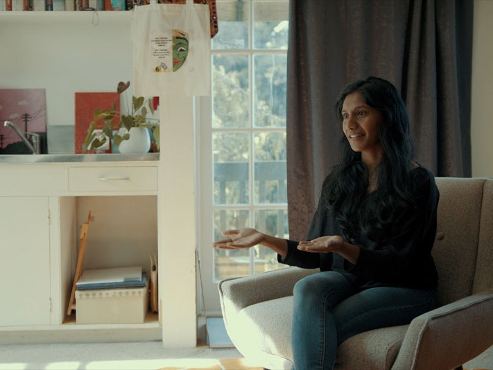 Still from ‘Healing doesn’t just happen in one mode’: reconsidering what ‘therapy’ looks like, 2023. Photo by Julie Zhu Video still of a woman sitting on a chair talking to somebody off-camera. Behind her is a window with curtain partially drawn, and a bench with lots of books on it