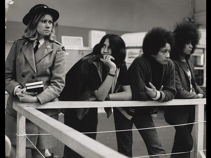 Ans Westra, Winter Show Building, Wellington, 1971. Purchased 1999 with New Zealand Lottery Grants Board funds. Te Papa (O.020448) View of three young Māori men leaning on a wooden rail. On their left stands a young blonde woman in a St Johns uniform.