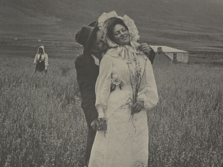 Jessie Buckland, Comin’ Thro’ the Rye, 1897/1908, Akaroa, silver gelatin print. Te Papa (O.045041) A man hugs a woman from behind in a field. She holds a bunch of flowers. Behind them a woman stands in isolation. Further back, there’s a stone house and hills