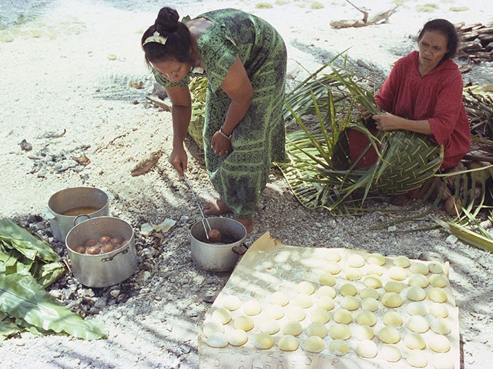 Glenn Jowitt, Woman cooking panikeke, 1981, Tokelau, colour negative. Gift of Glenn Jowitt Estate, 2015. Te Papa (E.007346) A woman prepares bread to fry in a pot, while another woman weaves with flax. They are sitting on a beach