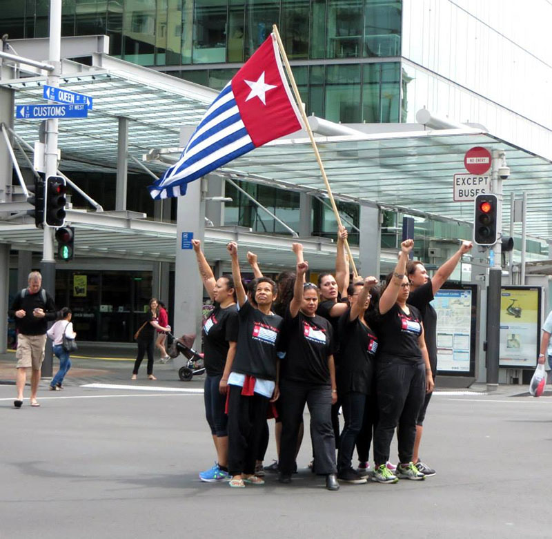 A group of women stand in the middle of an intersection with fists outstretched. One person raises the West Papuan flag