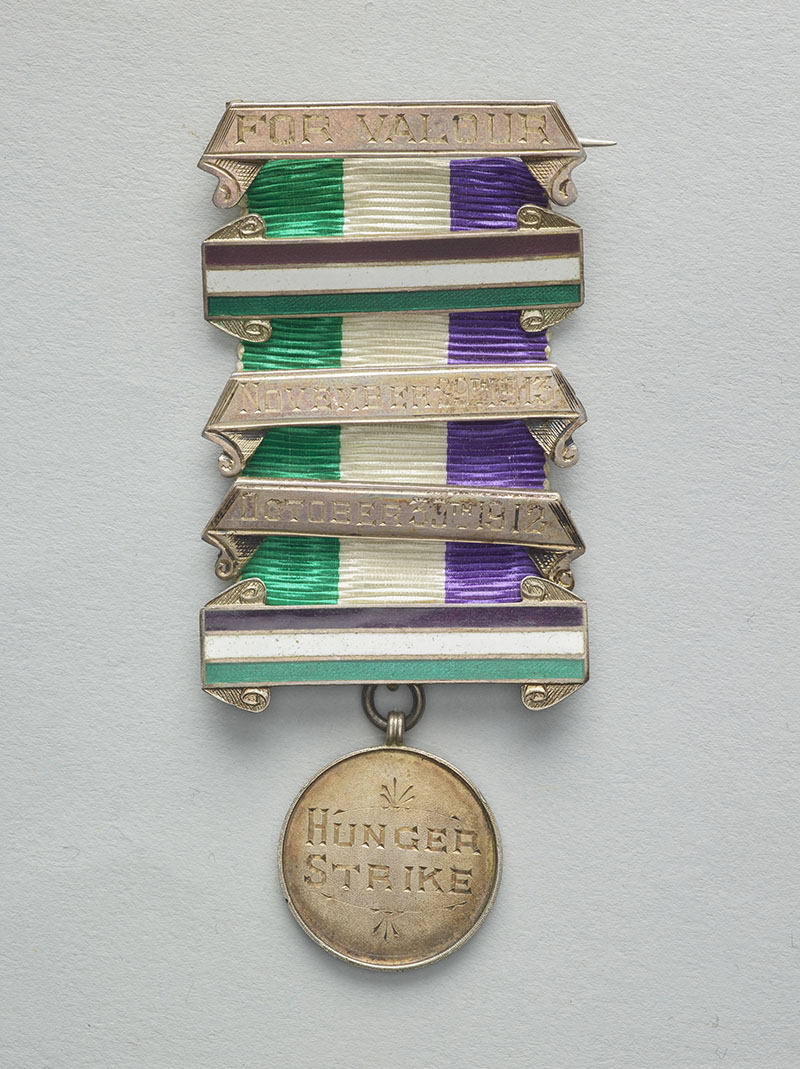 Medal, in two parts. The top part is a ribbon of green, white, and purple stripes contained by silver bands that say "For valour" and "November 29th 1913" and "October 30th 1912". Below hangs a circular piece with the words "Hunger strike" engraved in it