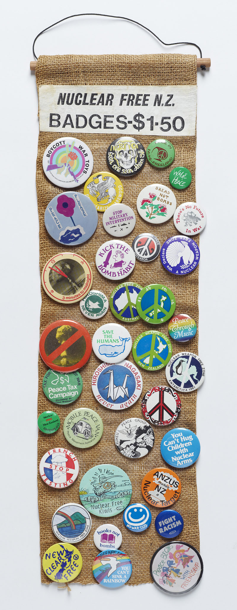 A piece of hessian cloth with a hanger and a sign saying "Nuclear free N.Z. badges – $1.50" on it. Attached to the hessian are dozens of vibrant anti-nuclear badges