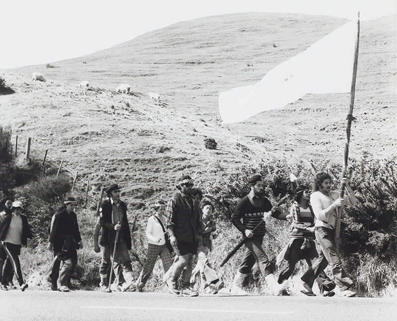 Māori Land March, 1975, by Ans Westra, gelatin silver print. Purchased 1993 with New Zealand Lottery Grants Board funds. Te Papa (O.009298/03) Black and white photo of a large group of people marching alongside a rural road. The person at the front is carrying a large flag