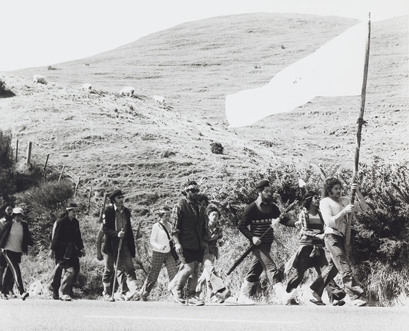 Black and white photo of a large group of people marching alongside a rural road. The person at the front is carrying a large flag
