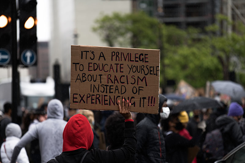 Photo of a crowded street full of protestors. One person, seen from behind, holds up a sign that says “It’s a privilege to educate yourself about racism instead of experiencing it!!”
