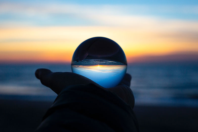 View of the early stages of a sunset over a beach. A hand holds up a glass sphere, inverting the scene