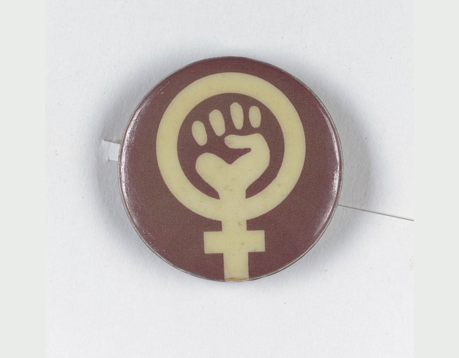 Brown badge with the female gender symbol on it. In the middle of the symbol is the silhouette of a fist