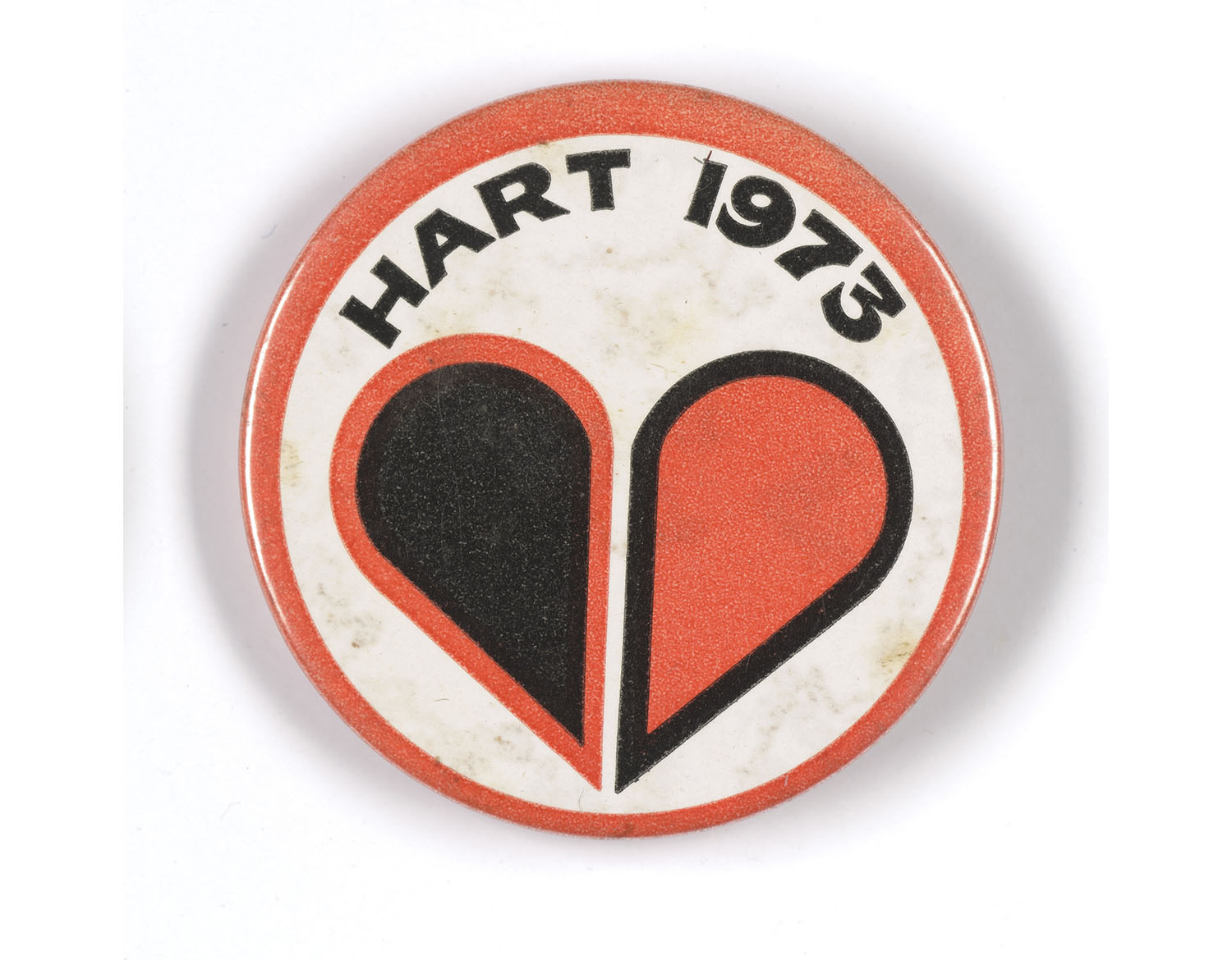Badge with a love heart on it. The heart is in two: one black with a red border and the other red with a black border. Above the heart are the words “HART 1973”. The badge has a red border