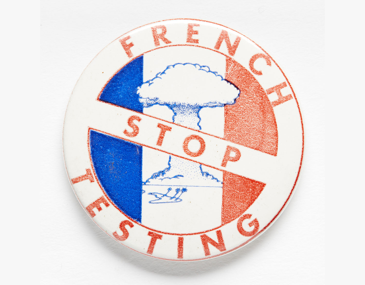 White badge with the French slide (blue, white, red stripes) on it. In the middle white stripe is an illustration of a nuclear bomb exploding. There is a strikethrough on this image with the text “Stop French testing”.