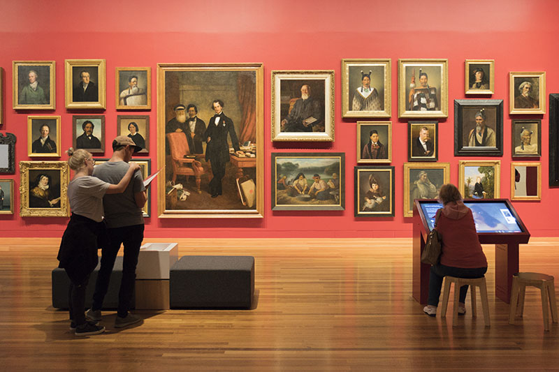View of the portrait gallery in Te Papa. On a red wall hang many portraits close together. In front, two people read an info sheet; beside them a woman uses a computer with an enlarged version of one of the paintings