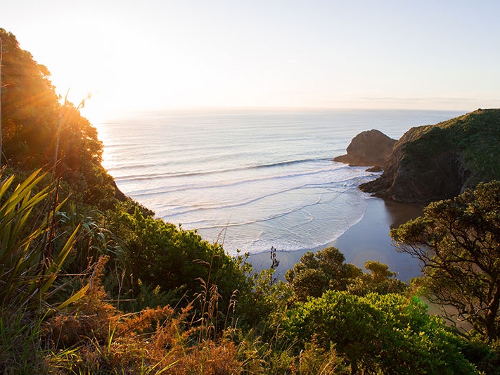 Whites Beach, Auckland, 2018. Photo by Kyle Myburgh / Unsplash View of a small cove and beach from above with the sun setting out on the water