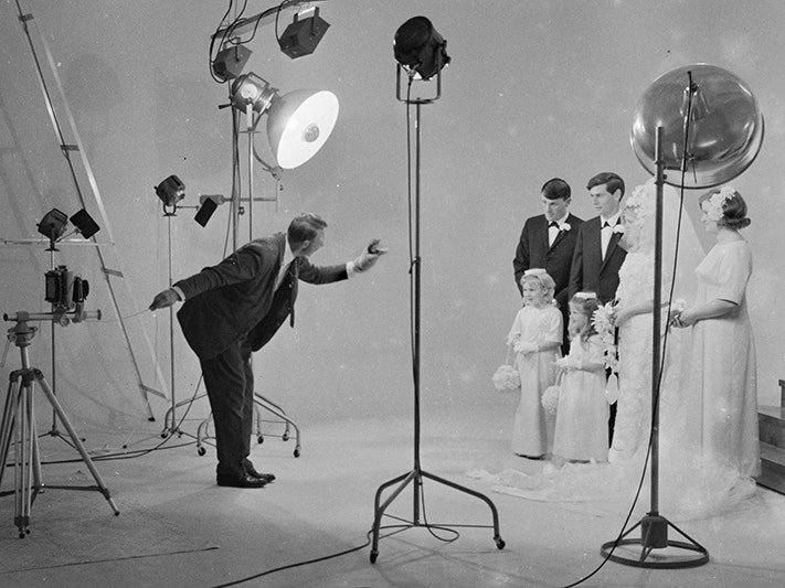 Crop of Christopher Bede Studio, by Frank Hofmann. Gift of the Frank Hofmann Estate, 2016. Te Papa (F.013805) A photographer has a puppet on one hand to entertain children while his other hand is operating a camera to take photos of a wedding party. The room is surrounded in lights and props.