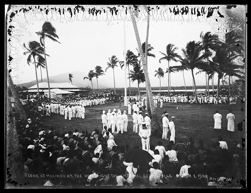 A black and white photo of many people gathered at an outdoor ceremony. There are palm trees and soldiers in white uniforms in the middle of the photo, and public looking at the ceremony from the sidelines.