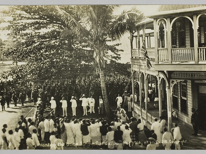 Postcard, 'Hoisting the Union Jack in Samoa. 30th Aug 1914.', 1914, Sāmoa, by Alfred James Tattersall. Purchased 2011. Te Papa (GH023108). A sepia photo of people in uniform in Sāmoa hoisting a flag - they are surrounded by troops and onlookers.