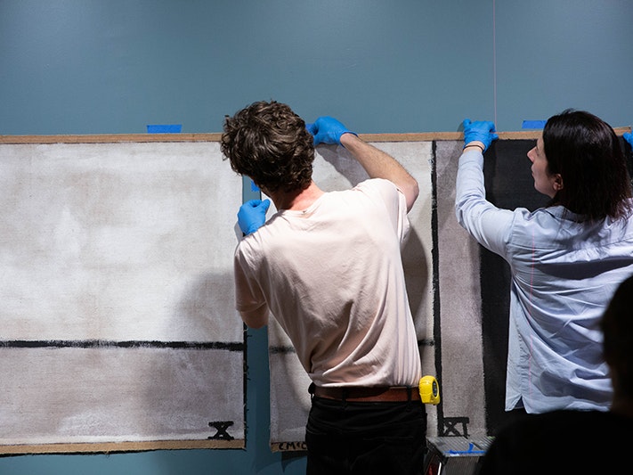 Te Papa staff installing Walk (Series C), 1973 by Colin McCahon, 2019. Photo by Jack Fisher. Te Papa (144200) Two people with their backs to the camera and wearing blue gloves are hanging a large painting on the wall.