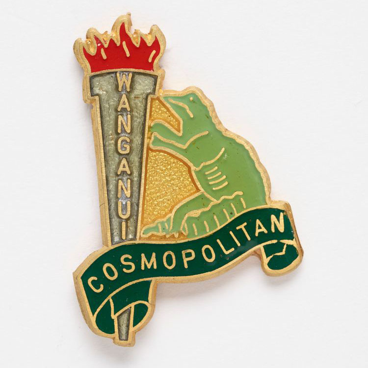 Decorative badge featuring a large flaming torch with the word Wanganui on it, and a green lizard standing beside it looking upwards at the flame. Underneath appears the word ‘cosmopolitan’