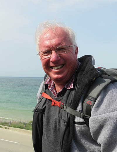 Head and shoulders of an older man standing near the sea. He is smiling at the camera and squinting into the sun.