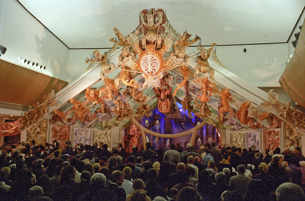 A photo of the back of a large crown in a big room with a sculptured stage at the end of it.