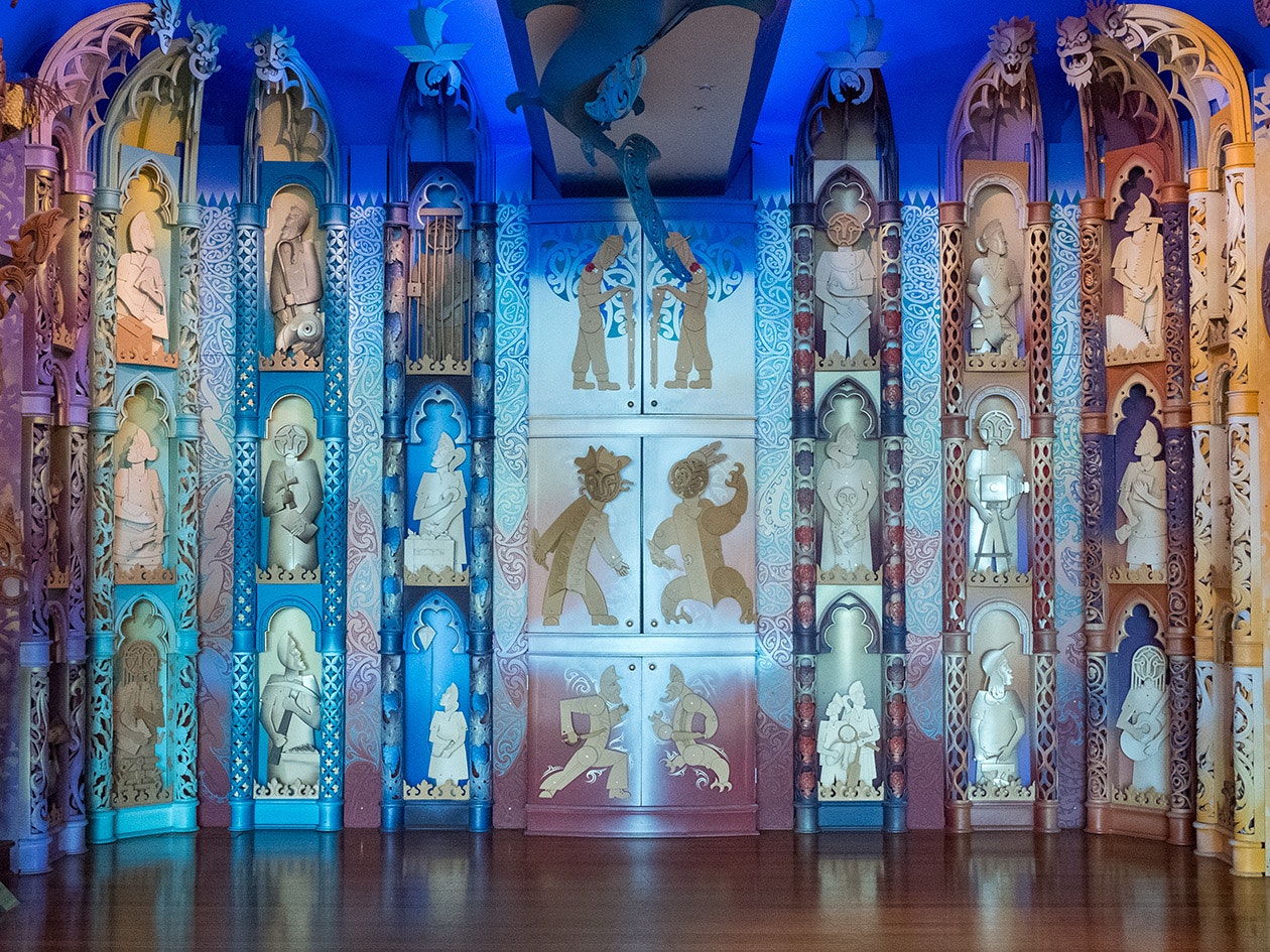 Detail of Te Hono ki Hawaiki, 2024. Photograph by Jane Harris. Te Papa  A colourful wall with carvings of stylised people doing different things and three cupboards in the central section.