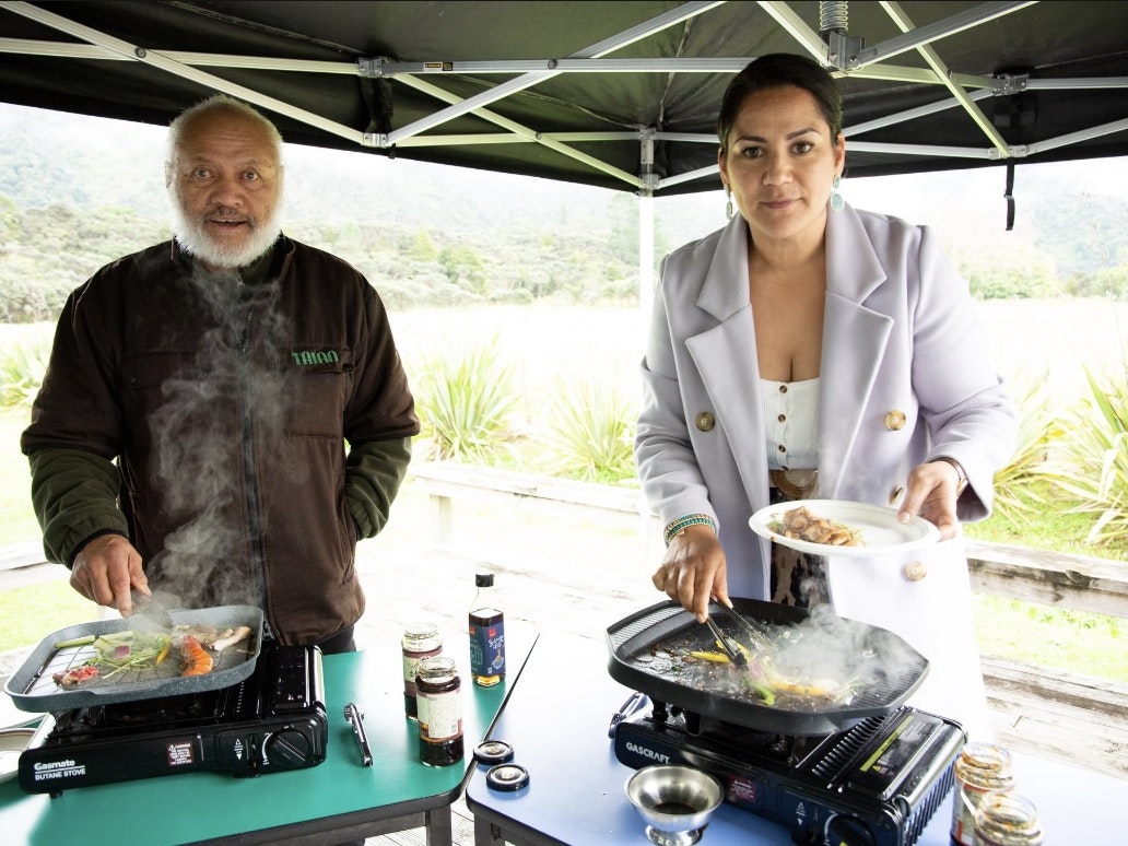 Rereata Makiha and Dallas King. Photo Courtesy of Dallas King Two people standing under an open marquee tent. Both are cooking on a frypan and looking at the camera.