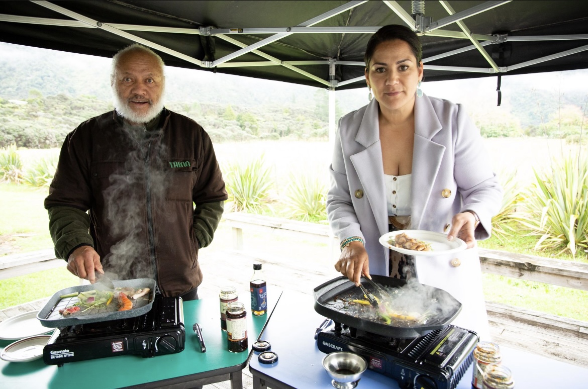 Two people standing under an open marquee tent. Both are cooking on a frypan and  looking at the camera.