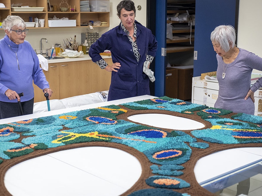 Joan Calvert, left, and Jean Ngan, right, with Conservator Textiles Anne Peranteau in the middle, admiring a panel from Forest in the sun, 2019. Photo by Daniel Crichton-Rouse. Te Papa Three women in different shades of purple are standing around a piece of carpet with large ovals in it sitting on a table.