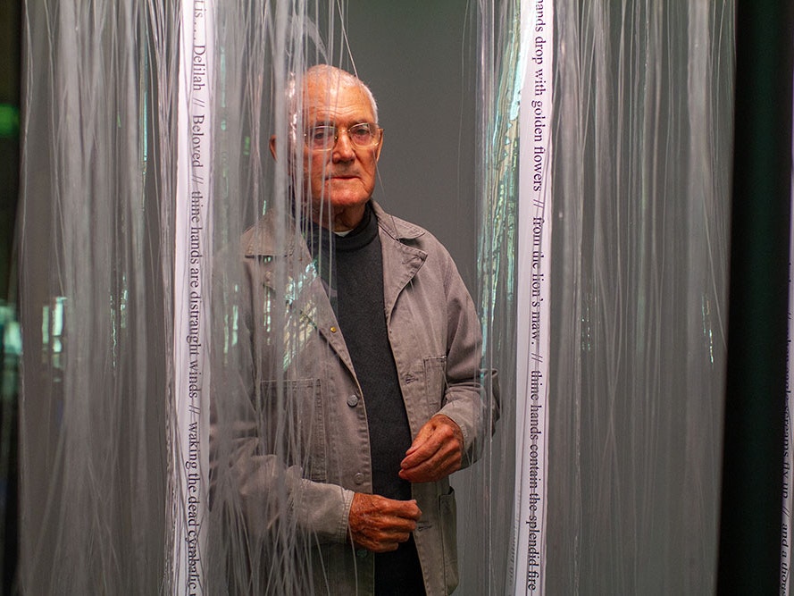 Jim Allen, Tribute to Hone Tuwhare, 1969, PVC, UV light, nylon thread, pegboard, square section aluminium tubing, vinyl, ink (2010 reconstruction). © Jim Allen. The poem Thine own hands have fashioned is reproduced courtesy of the Estate of Hone Tuwhare. Te Papa (2011-0014-1/A-I to I-I) A man standing behind transparent curtains.