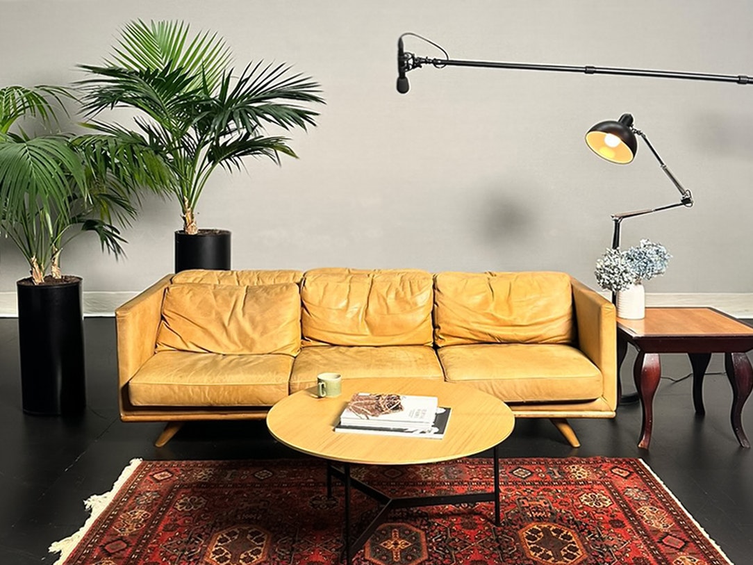 Interview set for manuhiri interviews, Not Your Dusky Maiden. Pua Flora 2023. Photo by Vioula Said. Te Papa A three-seater sofa with two plants on the left and a light and microphone on the right. There is a table in the middle.