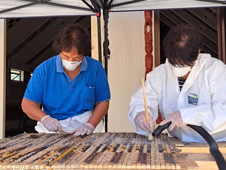 Aunty Elaine and Aunty Poppy brushing silt and dust off tukutuku panels. Photo by Migoto Eria Two women in masks are cleaning a woven panel damaged with mud.