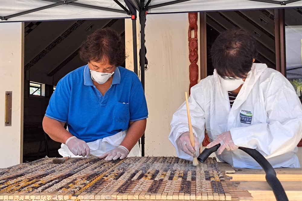 Two women in masks are cleaning a woven panel damaged with mud.