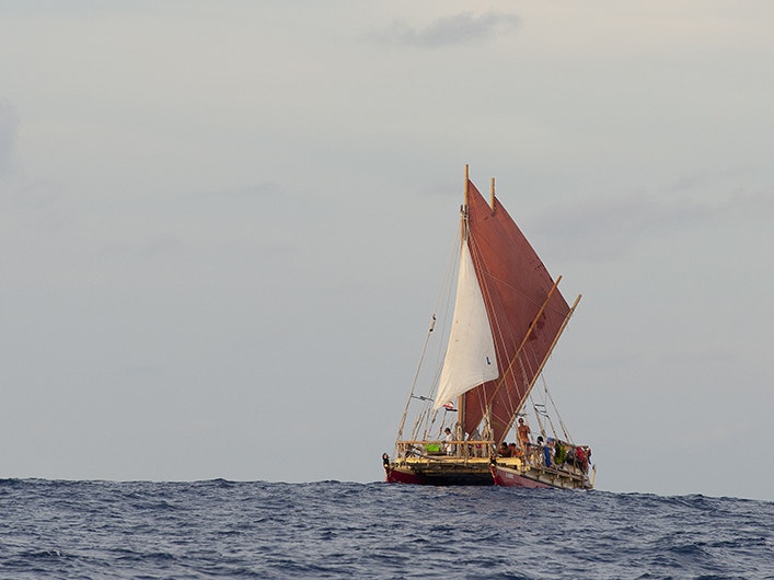 Te Aurere sailing in central Polynesia during the Waka Tapu voyage, 2012. Courtesy of Te Tai Tokerau Tārai Waka Inc A double-hulled boat is sailing on the sea. One sail is white the rest are red.