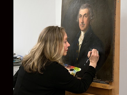 Jenny Sherman A woman is touching up an old painting on an easel in a studio.
