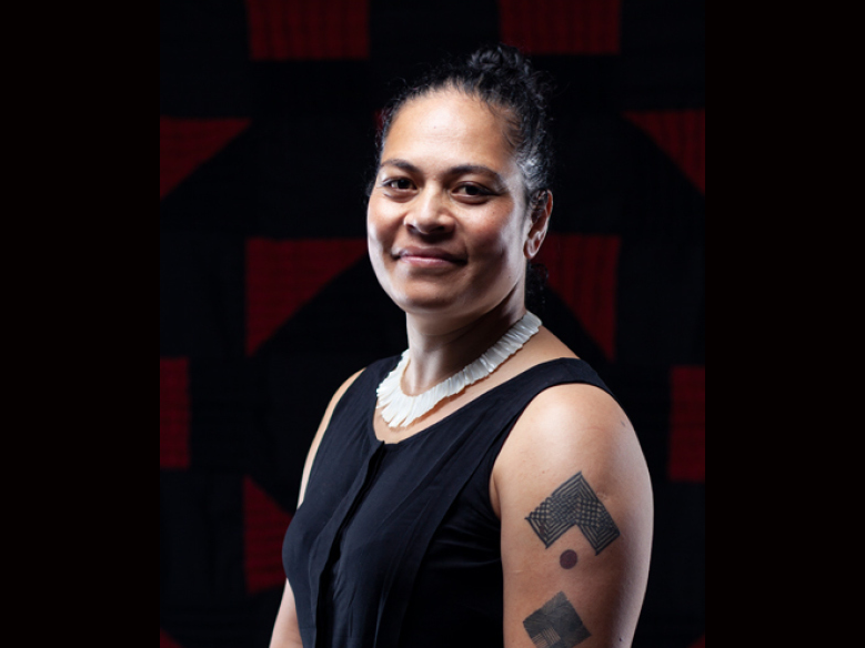 Kolokesa Māhina-Tuai Head and shoulders of a woman turned one-quarter to the camera. She has short sleeves and a tattoo on her arm
