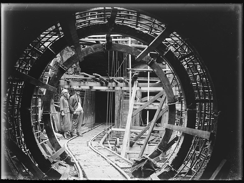 The bottom of the surge chamber viewed from the end of tunnel no. 2, and showing the head of the pipe line, 20 May 1923, Shannon, by Leslie Adkin. Gift of G. L. Adkin family estate, 1964. Te Papa (B.022979) A black and white photo of a man and a woman standing next to a railway track with a tunnel shape framing them.