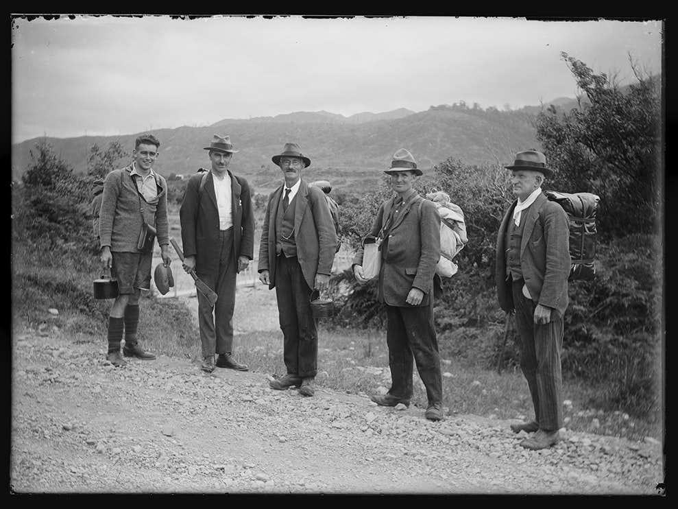 The "high country" party - Messrs Elsdon Clark, C. H. Martin, E. S. Lancaster, G. L. Adkin + J. Pollock - with packs up ready to start from Pipe Bridge on Gladstone Road, Ohau Valley, Levin. Black and white photo of five men in jackets and hats with hiking gear on their backs. They are all looking at the camera.