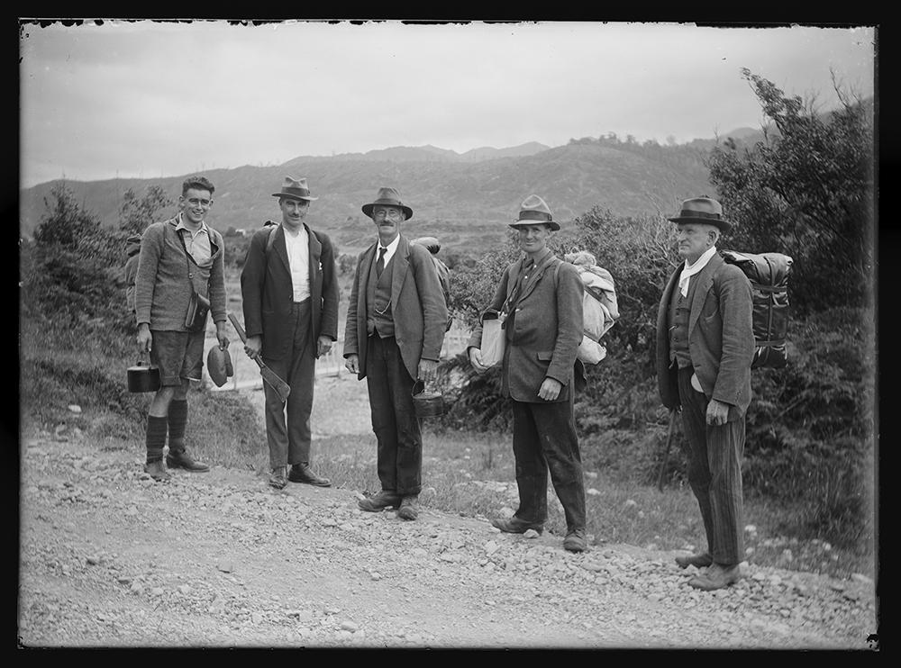 Black and white photo of five men in jackets and hats with hiking gear on their backs. They are all looking at the camera.