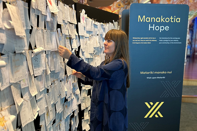 A woman in a long blue dress is tying a note to a wall of white paper notes.