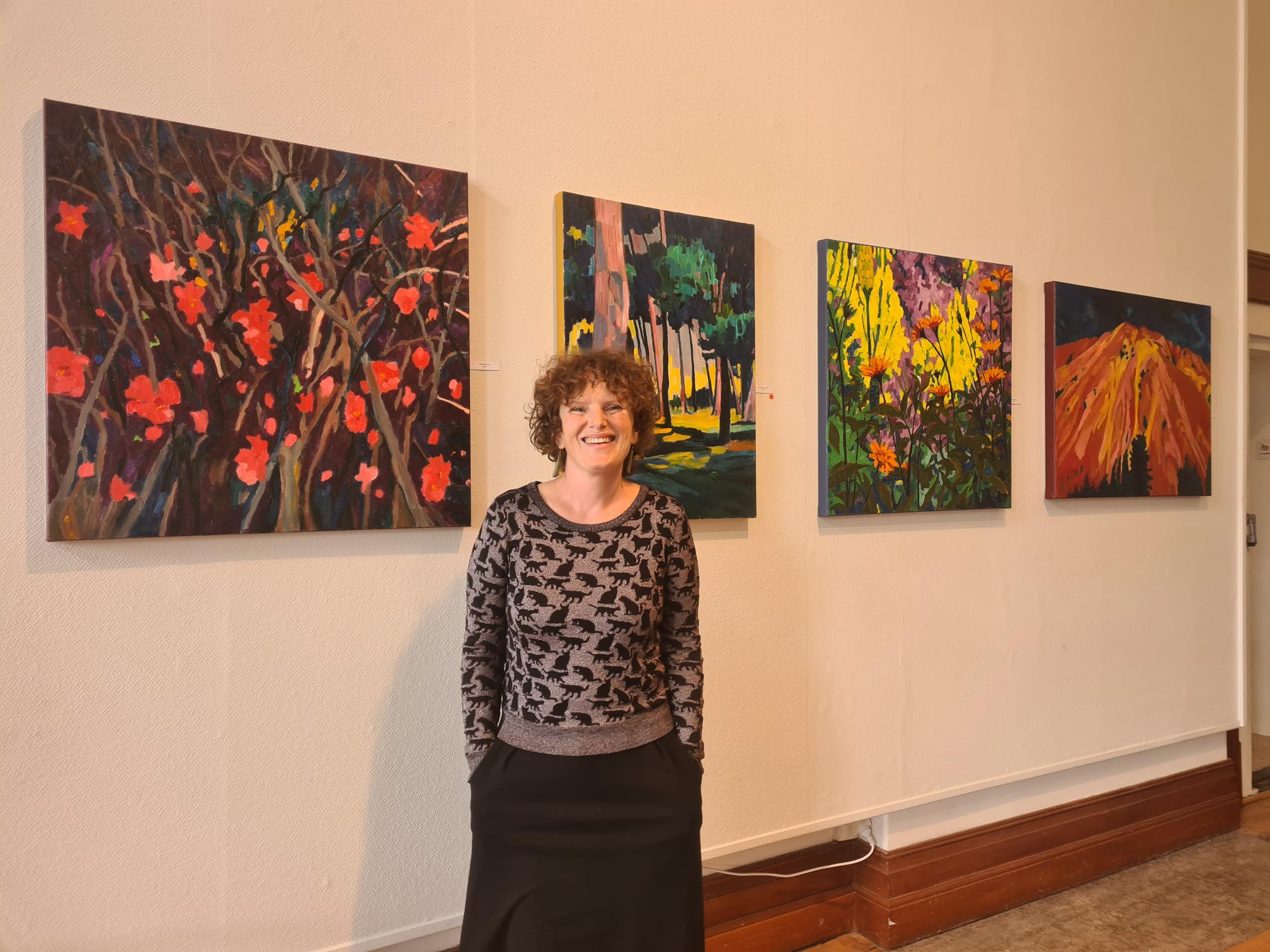 A woman is standing next to a wall that has four paintings hung on it.