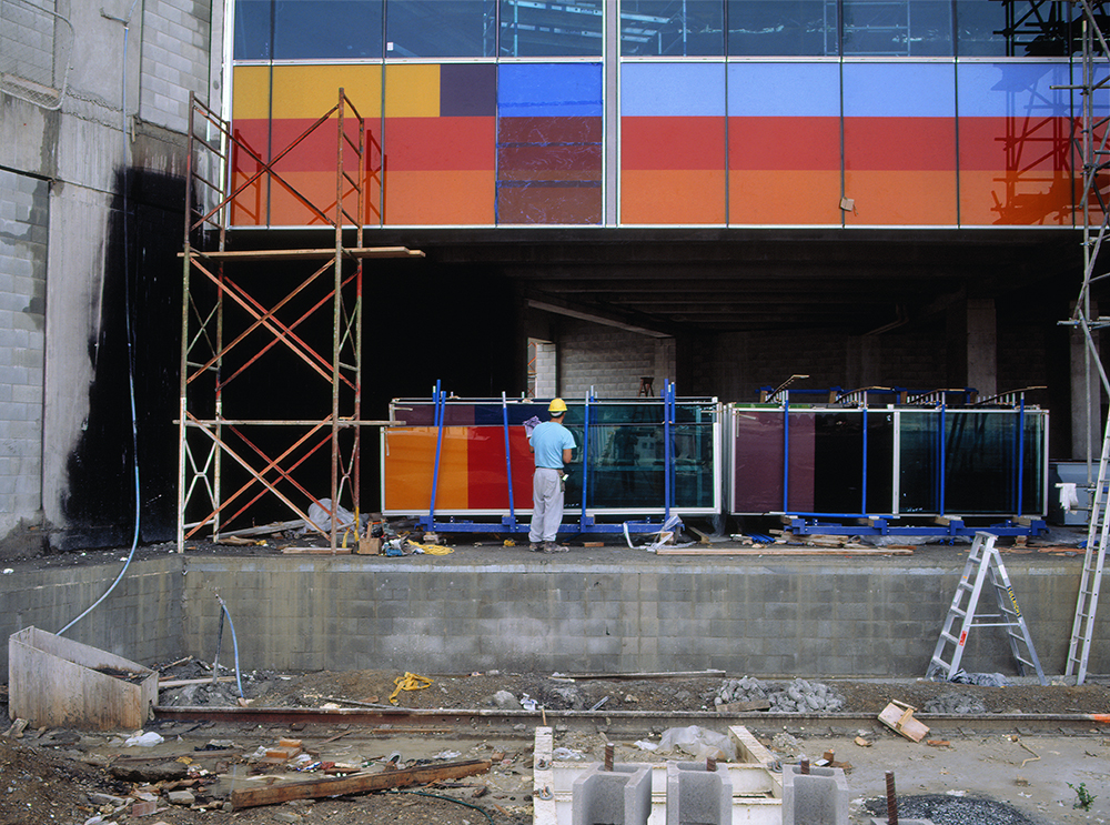 A man is holding a large glass panel in place under a larger wall where another piece of the glass panel is already installed.