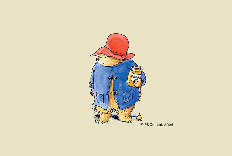 Illustration of Paddington Bear, wearing a blue trenchcoat and red hat, holds a marmalade sandwich