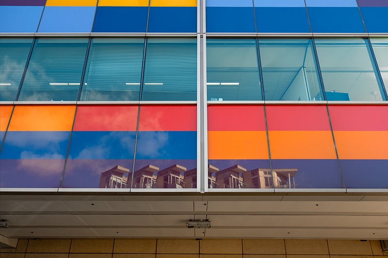 A wall of brightly coloured windows with a reflection of another building on the bottom windsows.