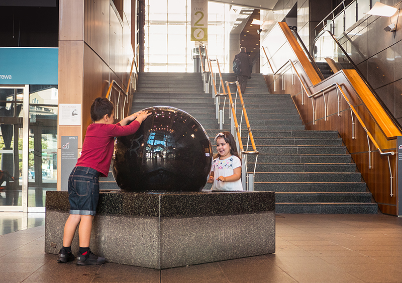 Two young children have their hands on a big black ball made of stone set in a stone box. The ball has water on it.