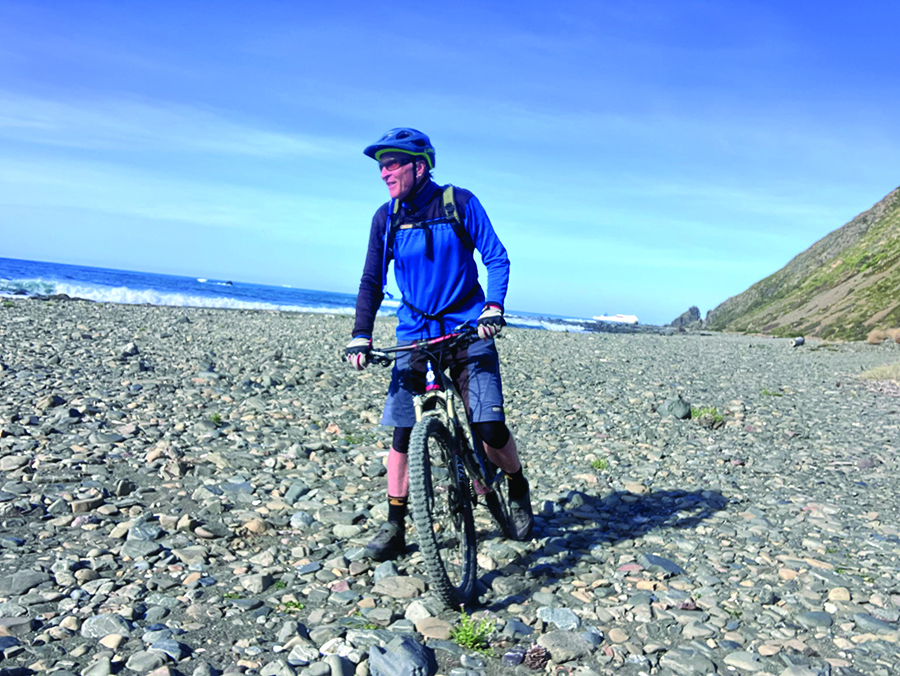A man in blue and black helmet and riding clothes is on a pushbike on a rocky beach.