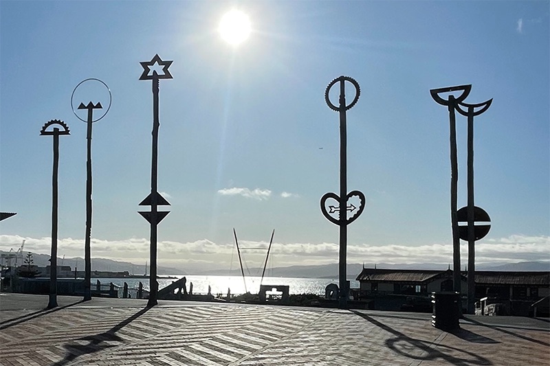An early morning photograph of the six sculptures on poles on the top of Wellington's City to Sea bridge. The harbour is in the background and the sun is also in the photo.