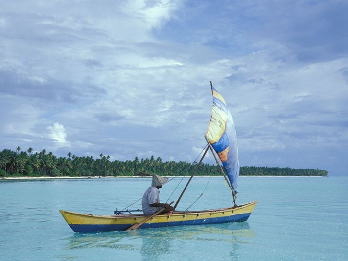 Tony Whincup, Untitled 4, Kiribati. Gift of Tony Whincup estate, 2017. Te Papa (O.047533) A man is in a small canoe that has a sail in front of part of a Pacific island beach.