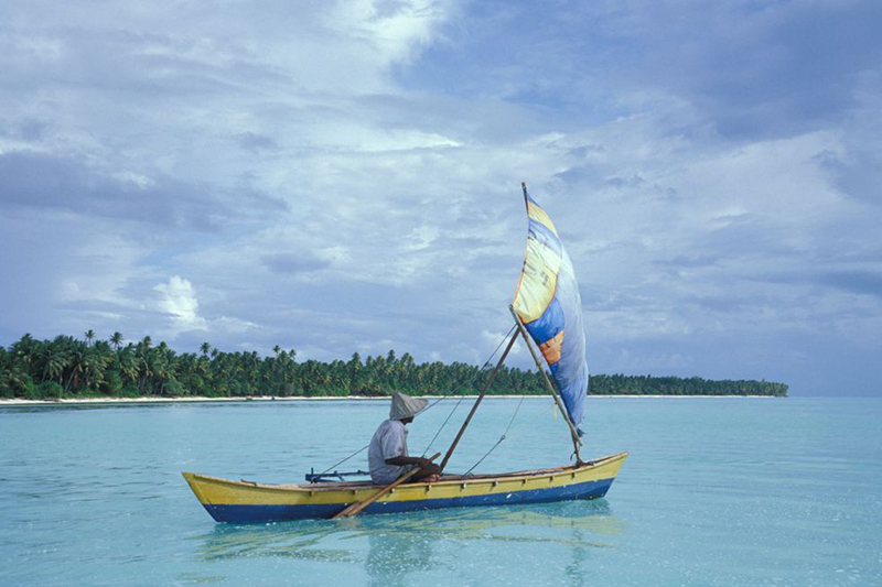 A man is in a small canoe that has a sail in front of part of a Pacific island beach.